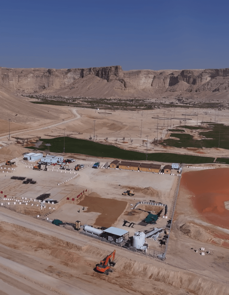 موقع بناء في وادٍ صحراوي يضم معدات ثقيلة ومباني مؤقتة تحت جبال صخرية شاهقة.