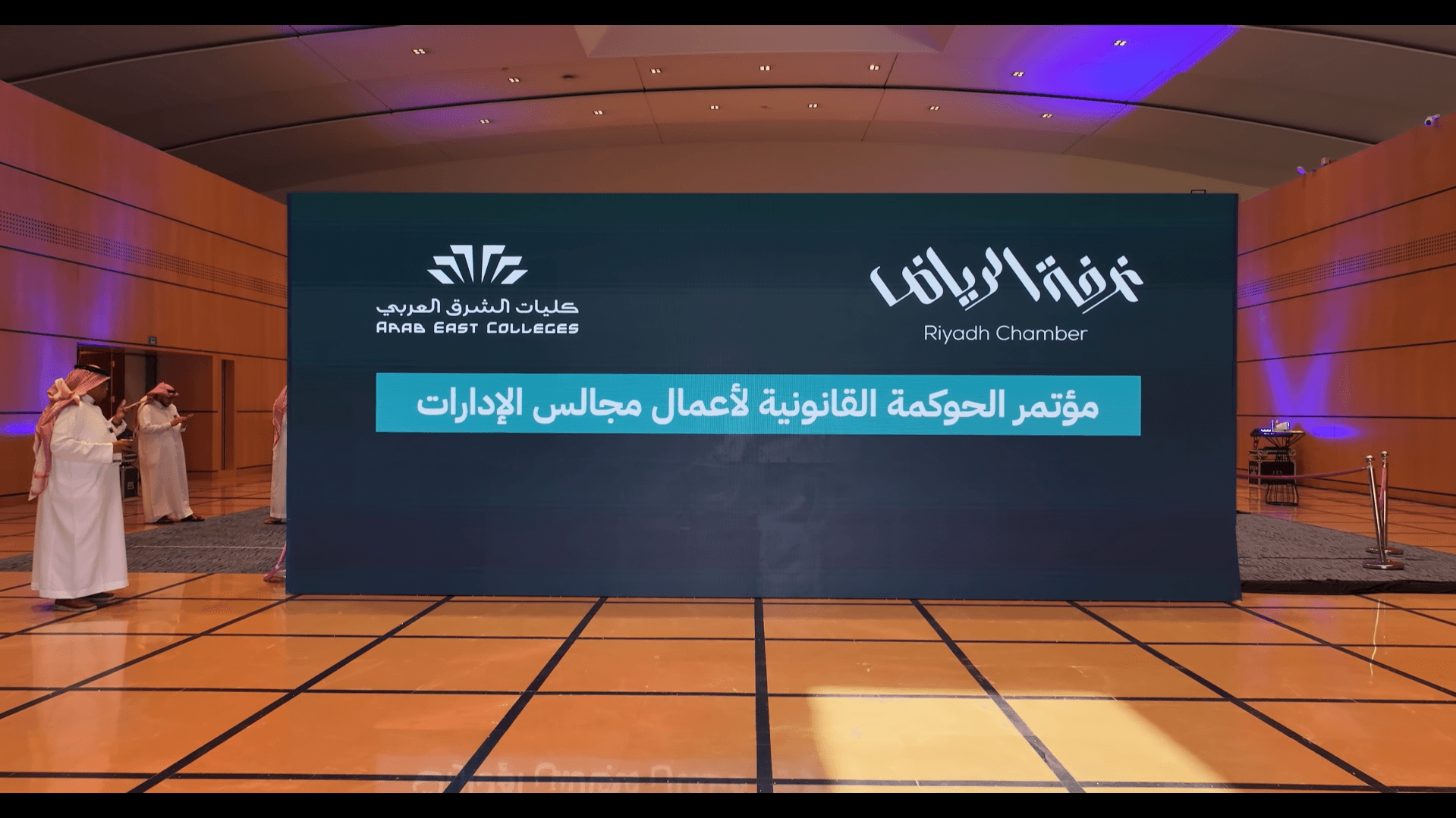 شاشة مؤتمر الحوكمة القانونية لأعمال مجالس الإدارات في قاعة واسعة بحضور أشخاص يرتدون الزي السعودي.