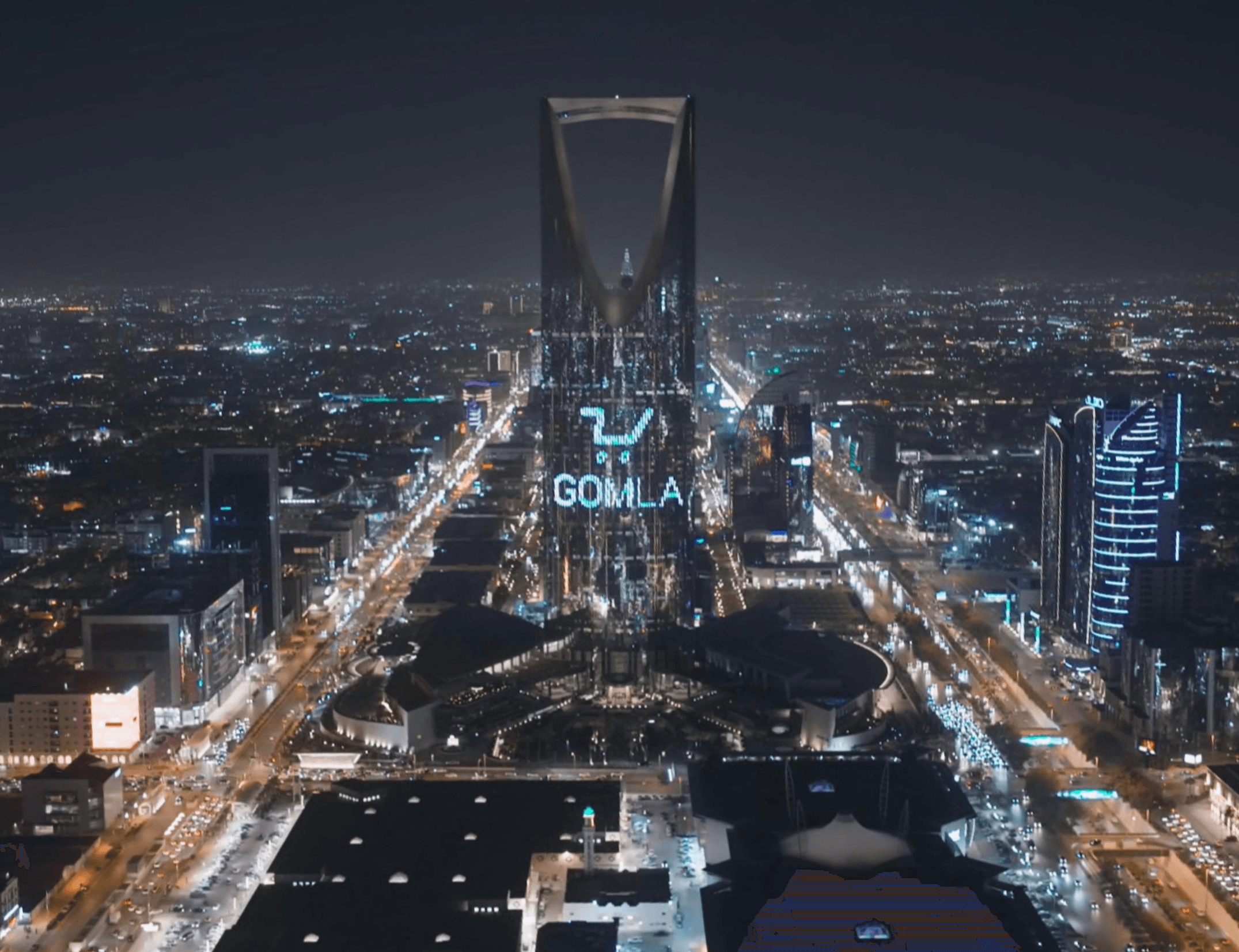 برج المملكة بالرياض ليلاً يعرض شعار "جملة" وعربة تسوق مضيئة فوق أضواء المدينة والشوارع المتلألئة.