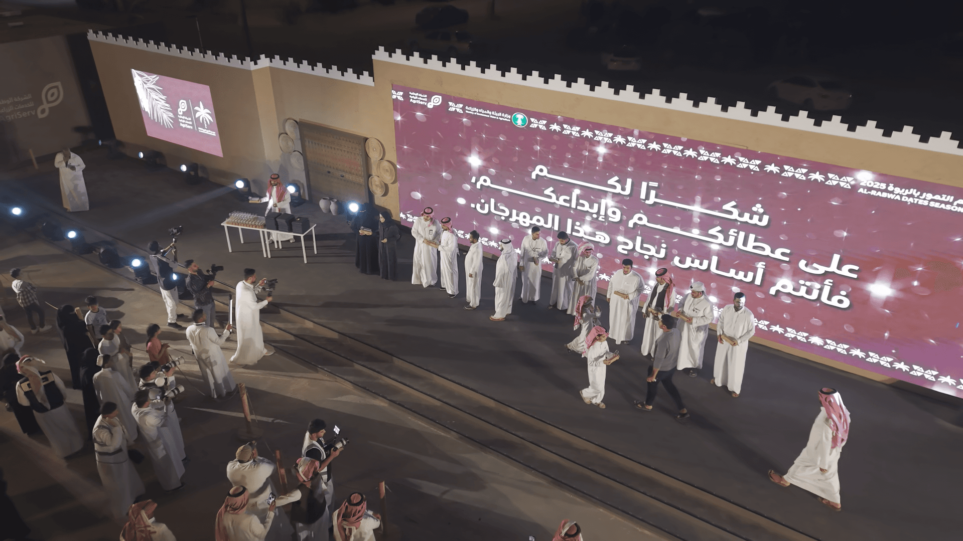 رجال يرتدون الزي السعودي التقليدي يقفون على منصة أمام شاشة كبيرة في مهرجان ليلي.