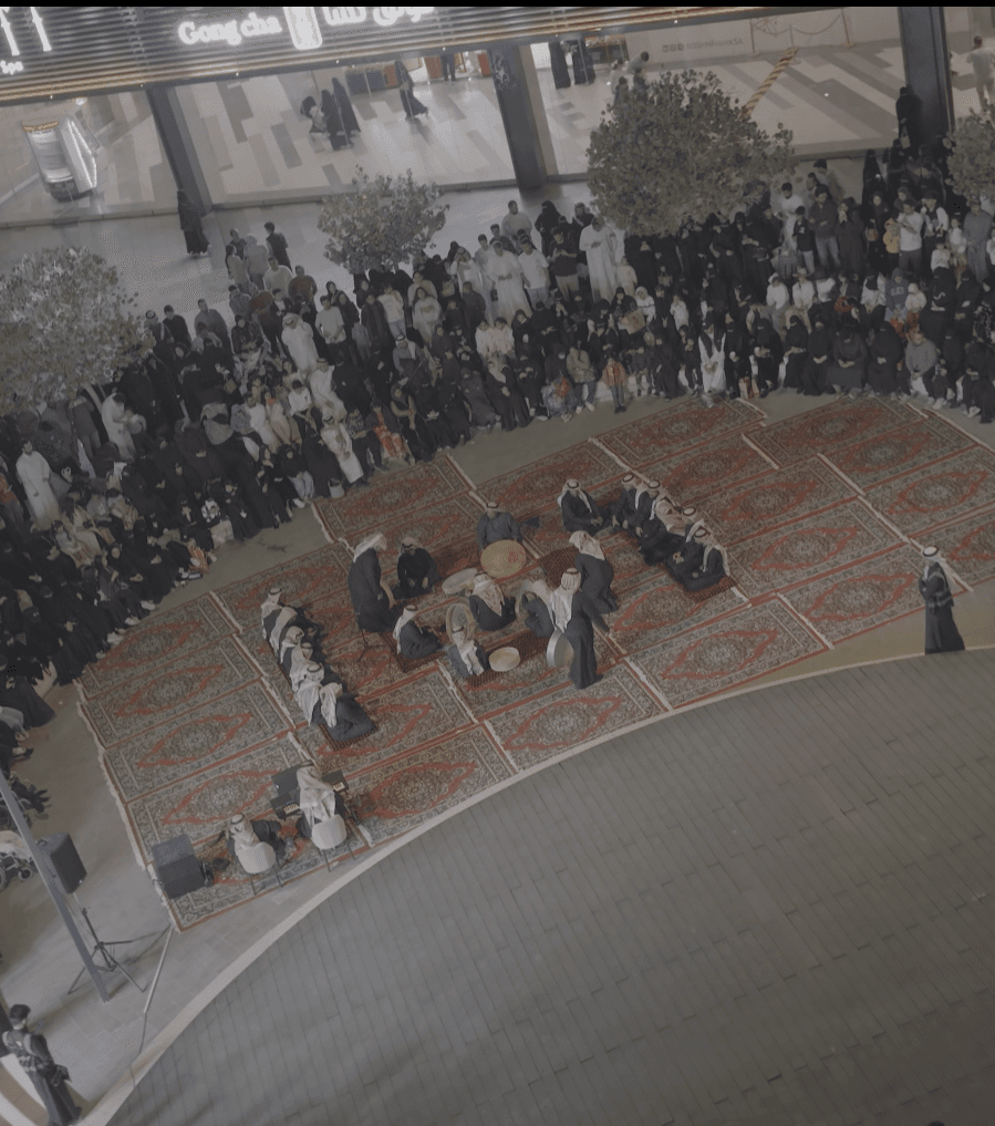 عرض فلكلوري سعودي على سجاد أحمر يحيط به جمهور كبير في ساحة مركز تجاري واسع.