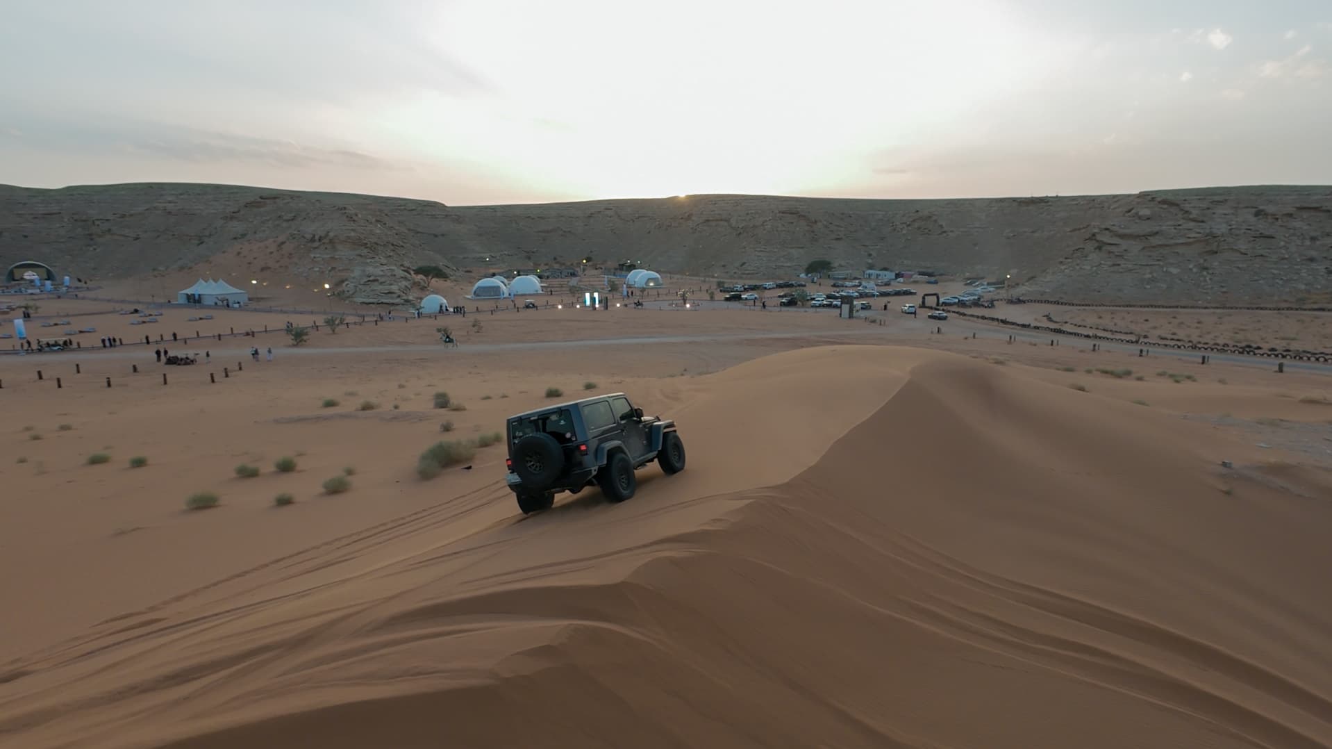 سيارة جيب رمادية تصعد كثيباً رملياً يطل على مخيم صحراوي به قباب بيضاء وقت الغروب.