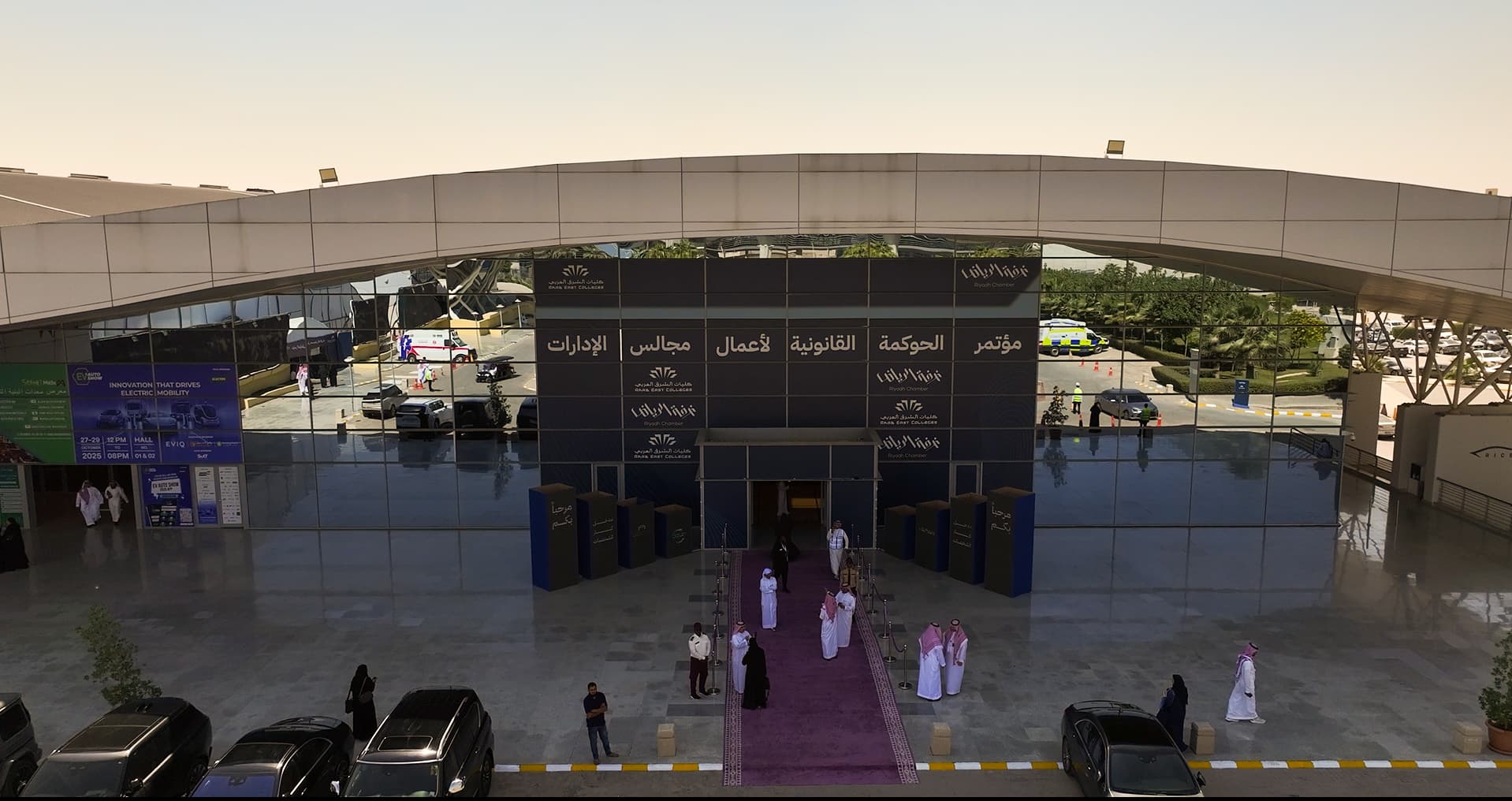 مدخل مبنى حديث بواجهة زجاجية وسجادة أرجوانية وأشخاص يرتدون الزي السعودي التقليدي في الرياض.