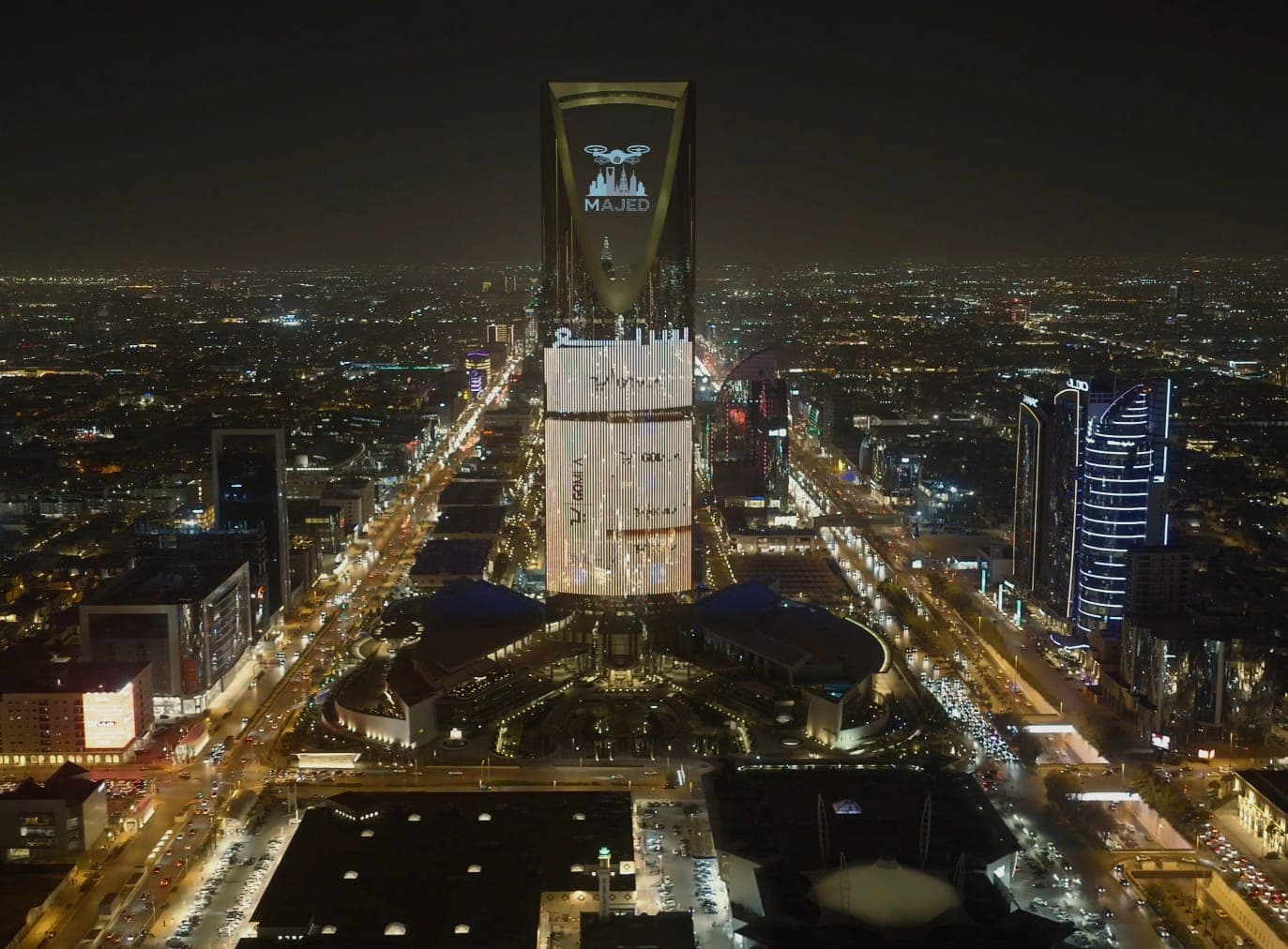 برج المملكة في الرياض ليلاً، مضاء بشعار ماجد وسط أضواء المدينة المتلألئة والشوارع المزدحمة.